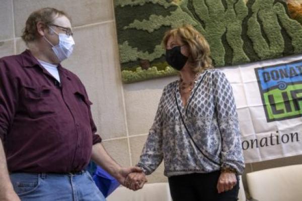 Dwayne Weller, who wears glasses and a face mask and is dressed in a chamois shirt and jeans, clasps hands with Maureen Stathes, who has shoulder length hair and wears a face mask, blouse and pants. A “Donate Life” banner is on the wall behind them.