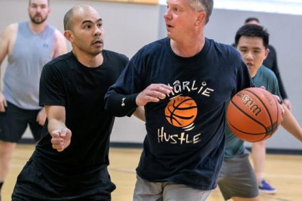Man wearing athletic clothing dribbles basketball up a basketball court as other men chase him and look on.