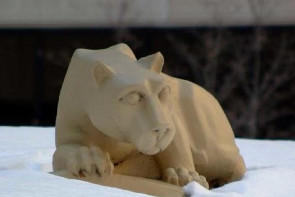 The Nittany Lion statue in the Hershey Medical Center courtyard is surrounded by snow, behind it, several trees and part of the Hershey Medical Center building are visible.