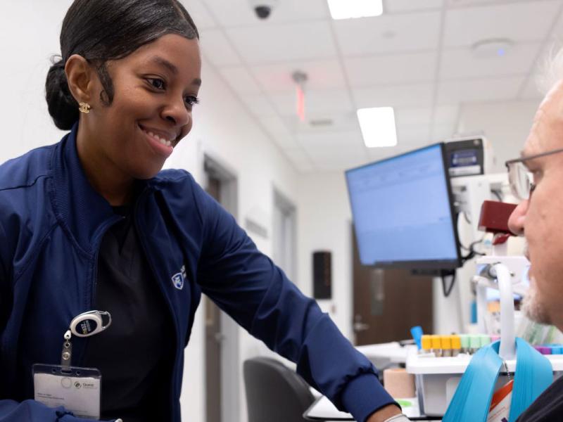 A female lead lab assistant is standing next to a seated male patient in a hospital lab office.  She is holding his arm to find a vein as she prepares to take a blood sample.  The lab assistant is wearing a navy-blue uniform and gray surgical gloves.
