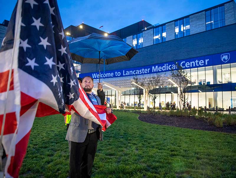 A man raises an American flag in front of Penn State Health Lancaster Medical Center. Behind him, a man holds up an umbrella. A sign on the front of the hospital reads “Welcome to Lancaster Medical Center.”