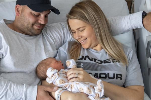 A happy couple sitting in a hospital bed, lovingly holding their newborn baby wrapped in a patterned onesie, with medical equipment visible in the background.
