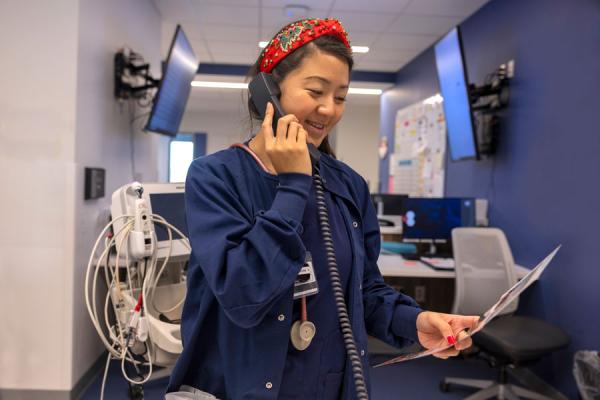 Women in blue scrubs on phone reading from a slip of paper standing inside a medical room.