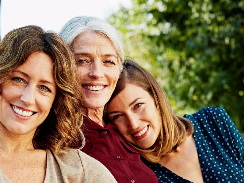 A mother and her two adult daughters are sitting happily together on a park bench.