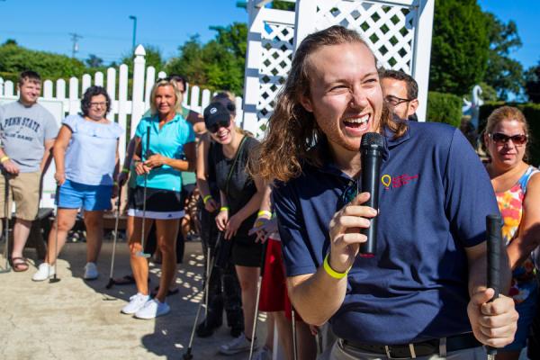 A young man stands at the front of a line, holding a microphone in one hand and a golf club in the other. Several people, who also are holding golf clubs, are lined up along the fence behind him.