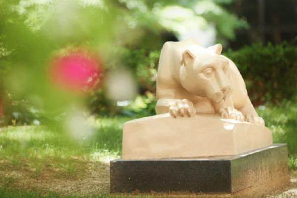 The Penn State Nittany Lion statue is seen in Penn State College of Medicine's outdoor courtyard. The statue is seen at the right of the photo in focus, framed by trees and pink flowers to the left and above and grass below.