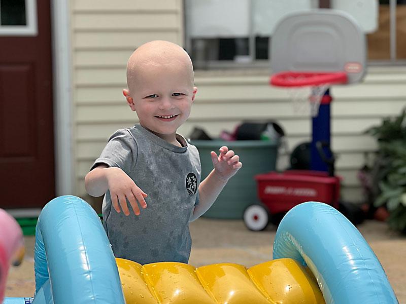 A young boy smiles while standing behind a backyard water slide. In the background, a box of toys and a children’s basketball hoop sit on the porch in front of a home. The door and windows are visible.