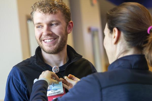 A smiling nurse wearing a badge labeled "Alexander, Registered Nurse" being assisted by a colleague who is attaching a pin to his uniform.