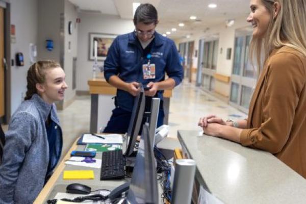 Three Penn State Health nurses engage in a discussion at a nurse's station in a hospital hallway.]