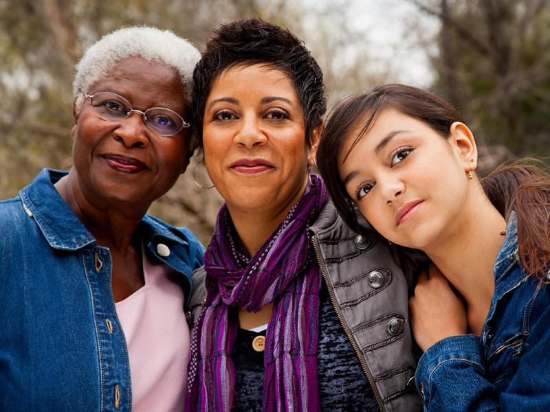 Three women stand close together, leaning their heads against each other. The woman on the left wears glasses. The woman in the middle has a scarf around her neck. The woman on the right has her hand on the shoulder of the woman in the middle.