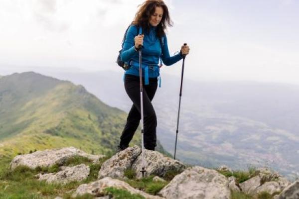 A middle-aged woman hikes in the mountains. She is walking on top of a mountain ridge, holding hiking poles, smiling and looking down.
