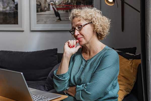 Senior woman with curly hair sitting on the sofa in the living room and using laptop.