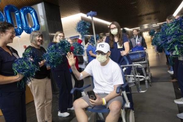 A man wearing a white t-shirt, white baseball cap, and face mask sits in a wheelchair and waves while holding a phone in his lap. He is surrounded by smiling hospital staff wearing scrubs and holding blue and green pom-poms.