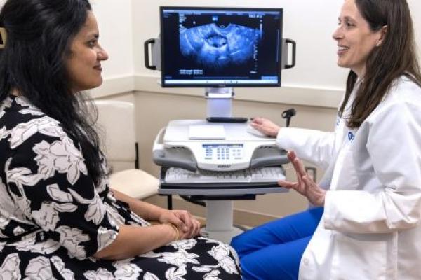 Dr. Sushrutha Sridhar, left, talks to Dr. Stephanie Estes of Penn State Health Obstetrics and Gynecology in a doctor’s office. Sridhar is wearing a flowered dress, and Estes is wearing a white lab coat. An ultrasound image of a uterus is shown on a screen between them.
