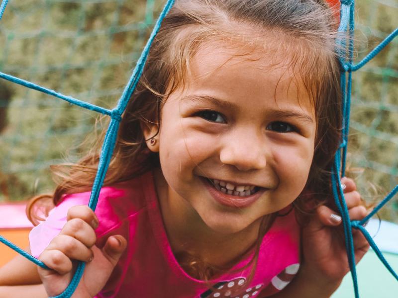 Little girl with pink shirt sits in a playground outside smiling at the camera.