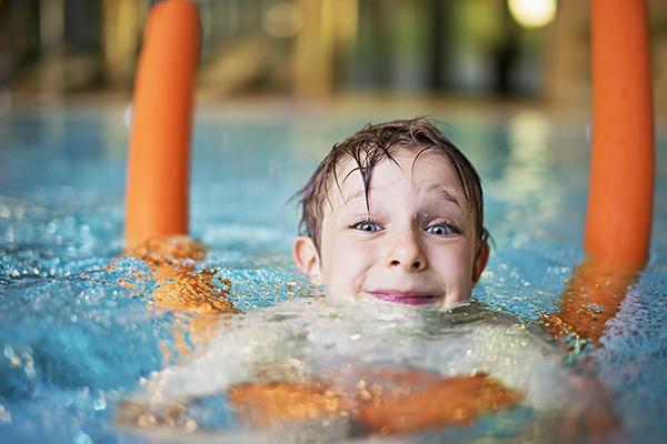 Happy kid learning to swim in indoors swimming pool. The boy is aged 5  and is using orange pool noodle. He is smiling into the camera.