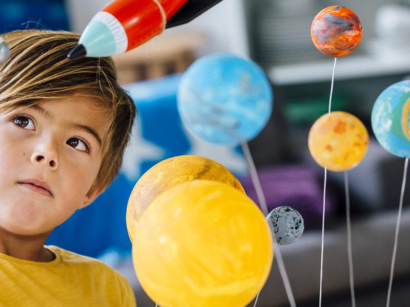 Little boy playing with his homemade planetarium as he holds an astronaut. A rocket hangs above. Arms raised as he plays.