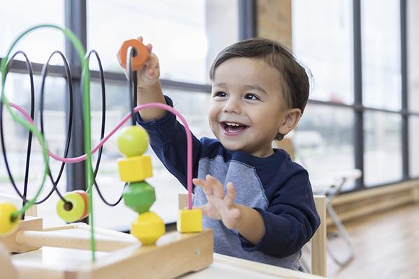 An adorable toddler boy sits at a table in a doctor's waiting room and  reaches up cheerfully to play with a toy bead maze.
