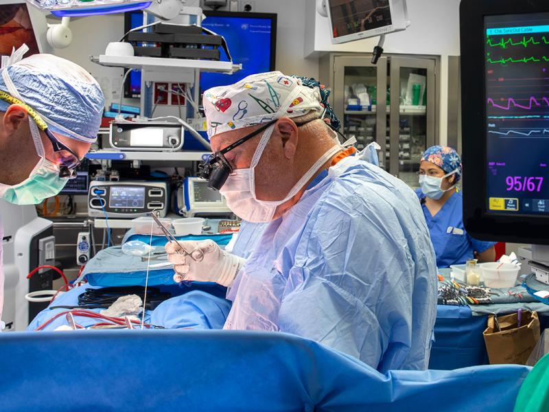 Two surgeons perform congenital heart surgery. They are dressed in scrubs, face masks and protective eye wear that also magnifies the surgical area. A nurse dressed in scrubs and a face mask is visible on the right. The background of the photo is filled with high-end monitors, surgical tools and supplies.