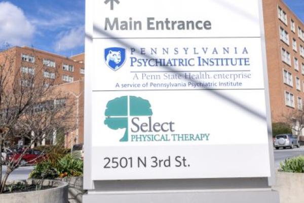 A brick, six-floor hospital is in the background. A sign in the foreground includes the logos for Pennsylvania Psychiatric Institute and Select Physical Therapy.