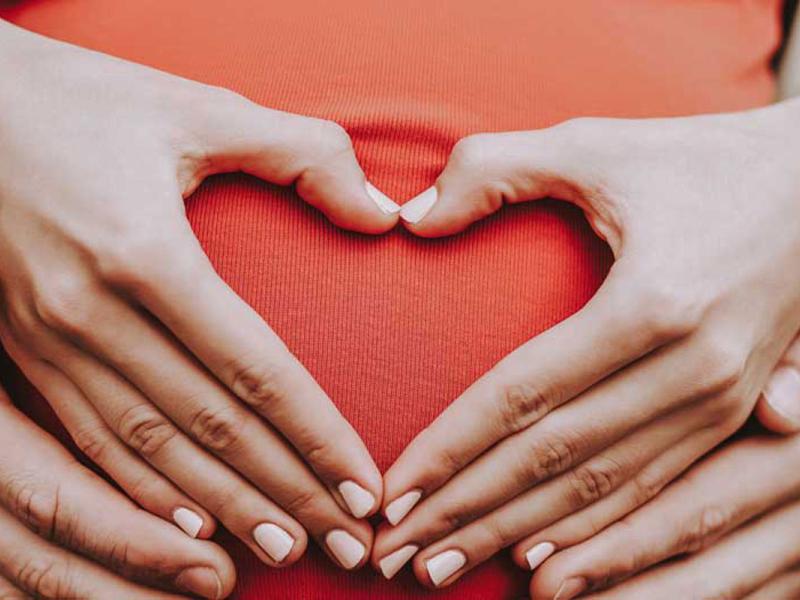 Pregnant woman sitting in front of a male with their hands on her belly in the shape of a heart.