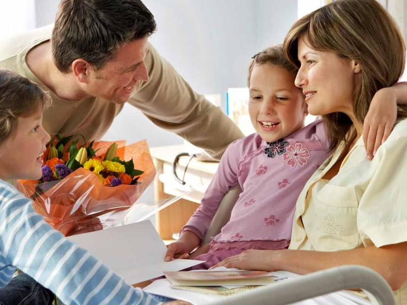 A smiling woman sits upright in a hospital bed while talking to her husband and two children.
