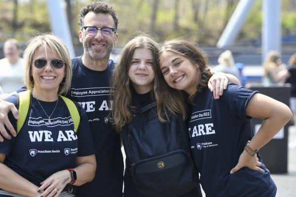 A smiling group of four individuals wearing "WE ARE" shirts at an outdoor Penn State Health Employee Appreciation Day event, posing together with arms around each other.