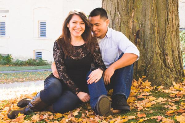 Man and woman smile at camera as they lean in to each other while seated against a tree.