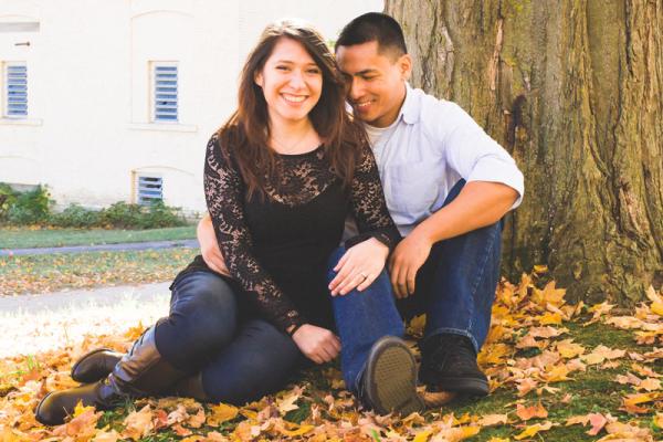 Man and woman smile at camera as they lean in to each other while seated underneath a tree.
