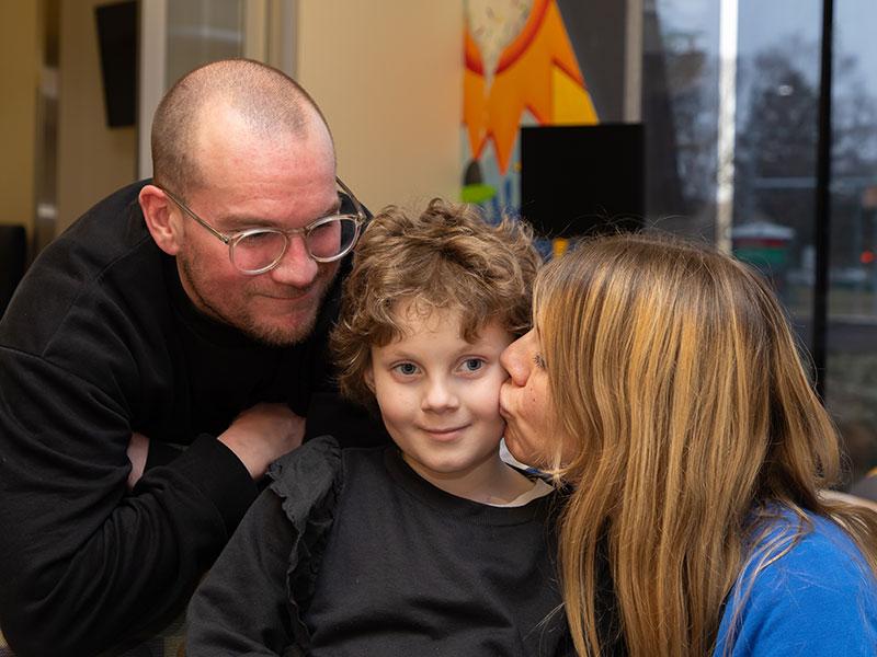 A man wearing glasses leans over the back of a chair while looking at woman kiss a little girl’s cheek. A window and monitor are visible in the background.