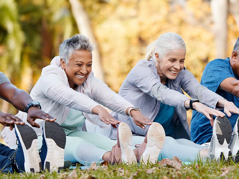 A group of four older adults, two men and two women, wear workout clothes as they sit on mats outdoors and reach for their toes to stretch.