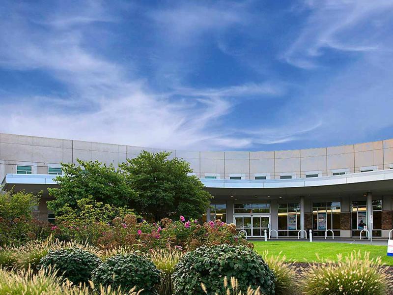 A driveway curves from the right of the photo to the front entrance of a hospital. Thin clouds fill the sky. A bell tower rises above Penn State Health St. Joseph Medical Center on the left. In the foreground is a garden filled with shrubs in front and trees along the back.