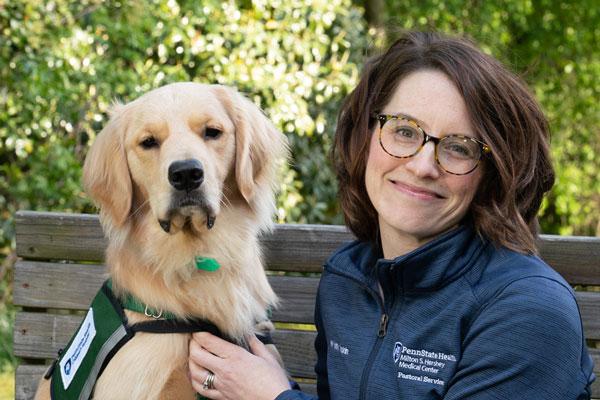 A golden retriever dog named Skye is on the left and a women wearing a navy blue top is on the left. They are sitting on a wooden bench with trees in the background. Both are looking at the camera. This photo was taken in April 2023.