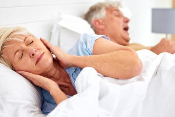 An older woman has her hands over her ears as her husband lies next to her snoring.