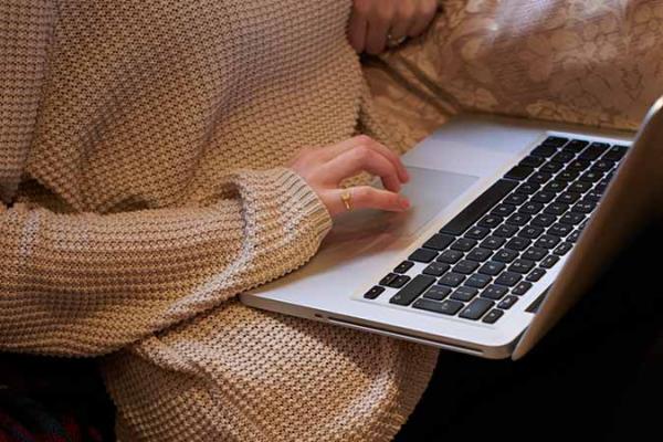 Closely cropped view of a person at home, sitting on sofa using a laptop that is positioned on their lap.
