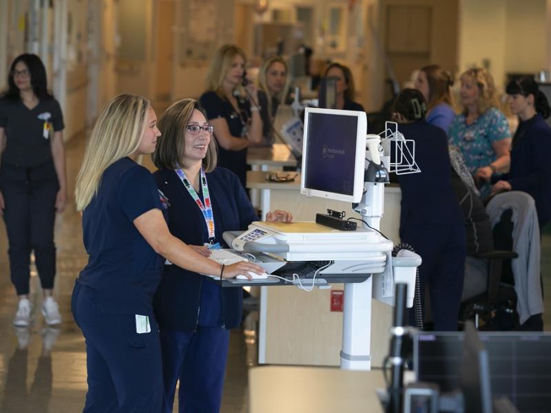 Two female nurses stand next to each other in front of a mobile computer cart. Both nurses are wearing navy blue scrubs. A group of nurses gather around a nurse's station in the background.