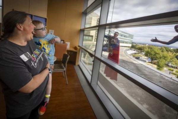 A woman holds a young child in a hospital room as they smile and look out the window. Outside, two window washers dressed as superheroes—Superman and Spider-Man—wave and gesture toward them.