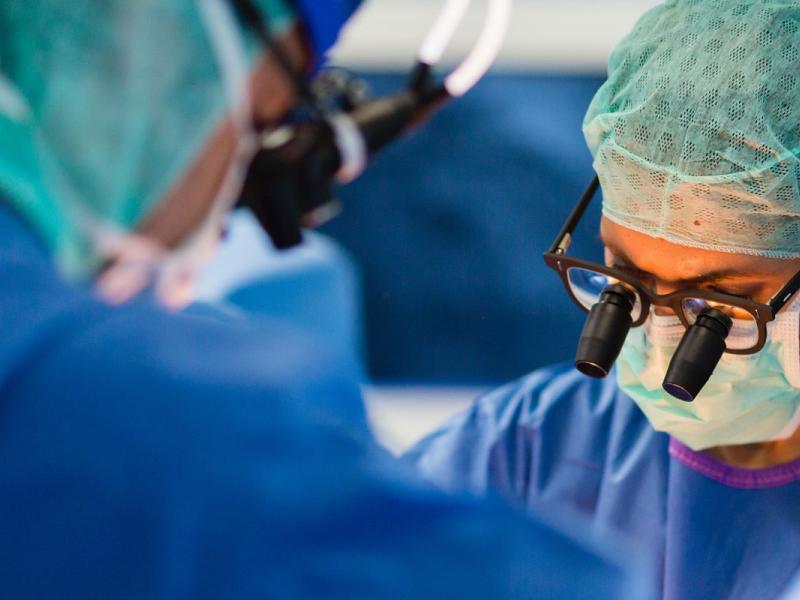 Two people dressed in surgical scrubs, including hats and masks, operate on a patient. They have microscopic surgical glasses strapped to their heads. Several monitors can be seen in the background.