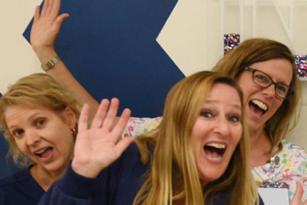 A group of nurses are seen in the Survivorship Clinic at Penn State Children's Hospital. They are making silly faces and gesturing to a large depiction of the word INSPIRE on the wall behind them.