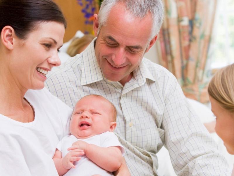 A woman sits in hospital bed with her newborn baby. Her husband and daughter look at the baby.