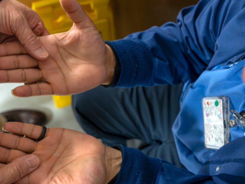 A close-up of two hands, one person assisting another by supporting their hands, with an individual wearing a blue uniform and an identification badge visible in the background