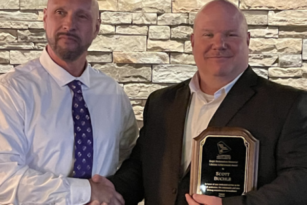 Two men shake hands in front of a stone wall. One man holds a plaque.