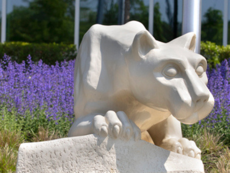 A white Nittany Lion statue crouching on a stone pedestal, surrounded by lush greenery and vibrant purple flowers, with a modern glass building reflecting the sky in the background.