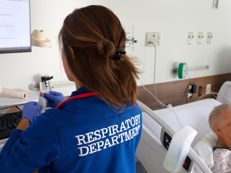 A respiratory therapist in a blue "Respiratory Department" jacket uses a computer workstation while monitoring an elderly patient receiving oxygen therapy in a hospital bed.